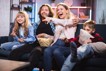 Caucasian parents with little son and daughter watching comedy on TV with toothy smile on faces. Happy family in casual wear sitting on couch and eating popcorn.