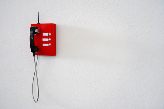 A red phone with a black handset and an antenna hangs on a white wall. Phone number for calling emergency services