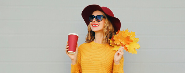 Autumn portrait of happy smiling young woman with yellow maple leaves and cup of coffee wearing a sweater and hat on gray background