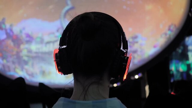 Woman wearing wireless black headphones and looking around in dark room of interactive exhibition or museum with colorful illumination - back view. Futuristic, immersive, entertainment concept