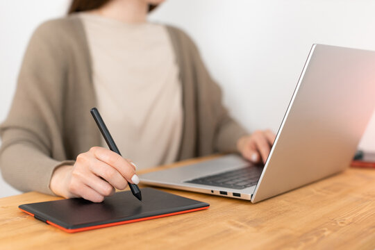 Unrecognizable girl in beige t-shirt and grey jacket sitting at wooden table opposite laptop and drawing picture on graphic tablet. Designer remotely work from home. Soft focus on hand with pencil.