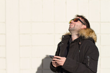 Fototapeta premium man laughs as he types a message on his smart phone outdoors, wearing sunglasses and a black jacket, selective focus