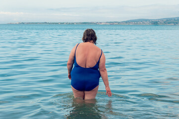 Rear view of overweight woman in blue swimsuit standing in still sea waters. Plus size woman at beach.