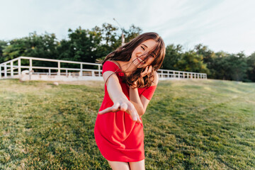 Fascinating brown-haired woman enjoying outdoor photoshoot in summer day. Joyful laughing female model in red dress standing on the green grass.