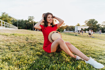 Positive white girl sitting on the grass and playing with her brown hair. Cheerful laughing lady in red dress enjoying weekend morning in park.