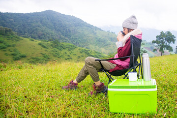Naklejka na ściany i meble Asian woman sitting on outdoor chair with drinking hot coffee and looking to beautiful nature of mountain. Beautiful female relax and enjoy outdoor lifestyle hiking and camping in summer vacation.