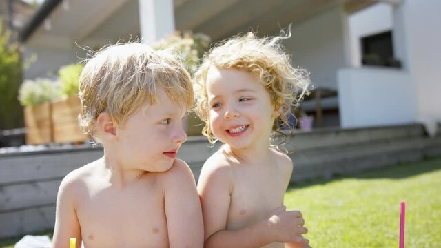 Front view of two sibling in swimsuit sitting next to pool and looking at camera.