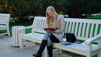 Fototapeta premium Young Beautiful Woman Sits On The Bench In Autumn Park And Works On The Tablet.