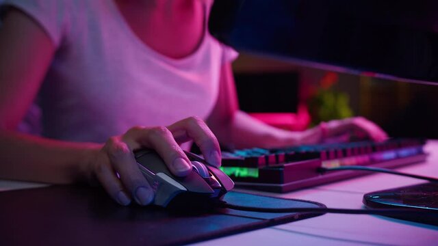 Woman gamer hands using wired computer mouse and typing on neon keyboard, playing video games at night close-up. Professional skills of cybersport. Gaming club concept. 