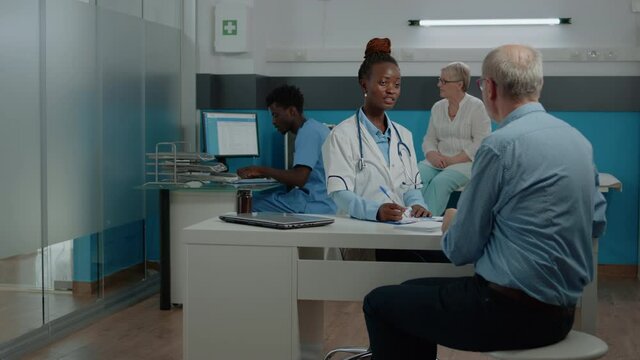 African american doctor examining x ray scan in hand to senior patient at desk. Black medic with radiography talking to elder man while nurse and old woman sitting in background