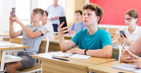Confident teen student using smartphone while studying in college with group of classmates..
