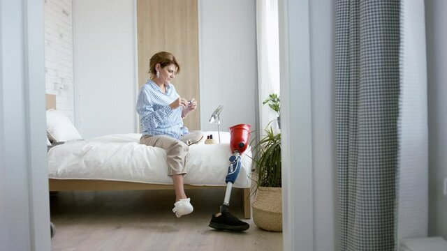Disabled mature woman sitting on bed indoors at home, looking at camera. Morning routine with leg amputee concept.