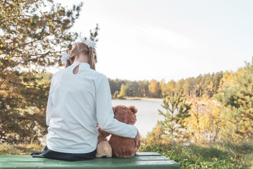 Happy little girl with stuffed toy teddy bear sitting on wooden pier near calm lake against autumn forest on sunny day back view