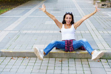 Fototapeta na wymiar Happy young fashion woman sitting on the street sidewalk stylish female model in a white t-shirt and in dark blue jeans arms wide apart from joy