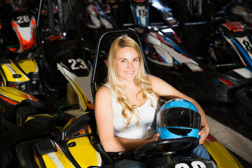 Fototapeta na wymiar Cheerful blonde young girl posing with helmet in her hands at kart circuit