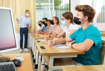 Teenagers in protective mask listening to lecturer and writing in notebooks in classroom