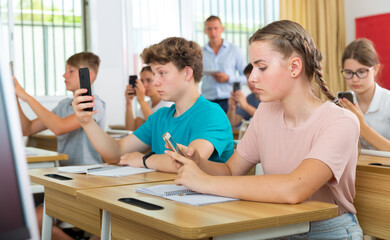 Group of modern teenagers sitting with mobile phones on lesson in classroom