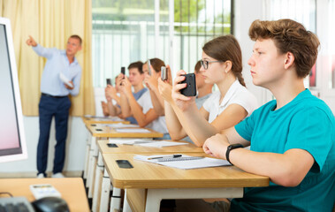 Confident teen student using smartphone while studying in college with group of classmates..