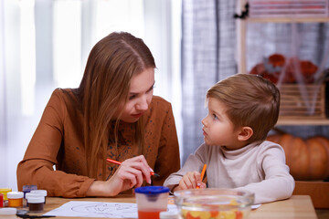 Mom helps her son draw a pumpkin with pencils. The family sits at the table with treats, in the...