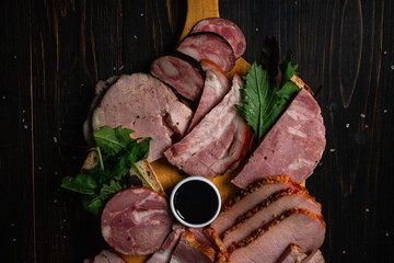 Slices of assorted sausages on a cutting board with dark sauce and green leaves. Dark wooden background. 