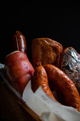 Assorted sausages stacked on baking paper in wooden box on black background.