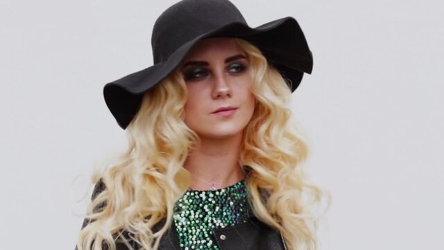 Stylish blonde girl in a black hat, black leather jacket and a shiny dress posing in front of the camera against a white wall background. Video