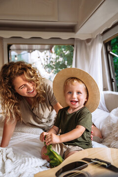 Joyful young mother enjoy being together with small kid boy. Portrait of little child in straw hat sit on bed next to curly loving female parent. Hippie carefree mom travelling with son in camper van