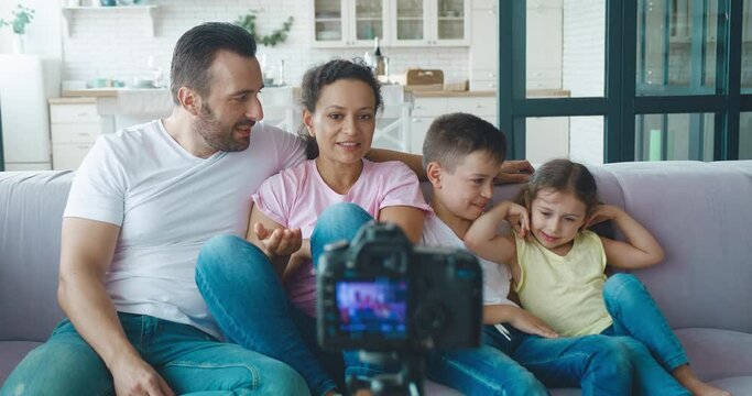 Multiracial family bloggers wave hello looking at the camera mounted on a tripod, actively gesticulating, smiling laughing, communicating and having fun during recording video blog