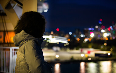 A slender young lady dressed in winter clothes standing on deck of the cruise ship and looking at Melbourne's city lights in the evening.-Portrait Photography.
