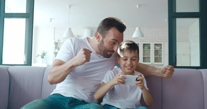 Cute boy playing games on smartphone and his excited father actively supporting him and gesturing with fist after win. Man gives high five to his son. 4K video