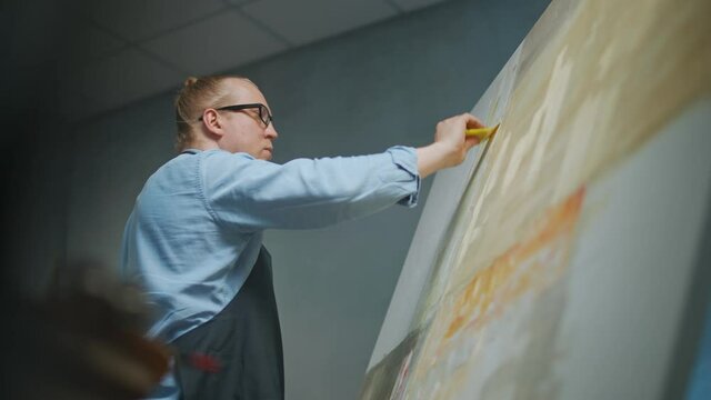 Artist's workshop, creative person at work, a talented adult man artist covers the canvas with beige oil paint, draws the modern abstract picture, low angle view, 4k slow motion.