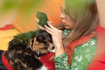 Young girl is sitting at home on couch with cat lying on her and with smartphone in her hands.