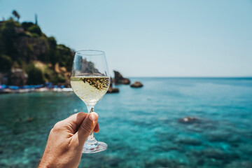 Glass of wine in hand. A glass of red wine against the background of the Mediterranean beach and the sea in a tourist town in the summer under sunlight. Summer, travel, lifestyle, relaxation, and