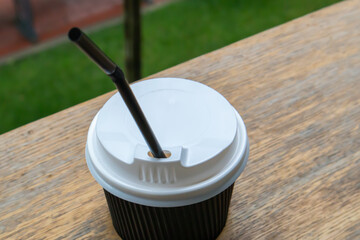 One black paper coffee cup with lid outdoors in summer in sunny weather on a wooden table of a cafe, coffee shop or restaurant.