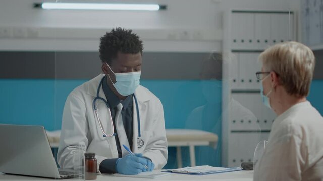 Medic Consulting Elder Patient With Face Mask And Gloves For Coronavirus Protection At Healthcare Facility. Young Doctor Helping Senior Woman During Pandemic At Checkup Appointment