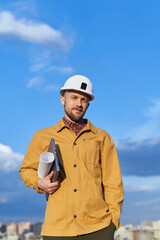 Man in white helmet and orange jacket standing on construction site. Cute caucasian bearded foreman, manager, architect or building contractor at sunny day with blue cloudy sky and urban big city view