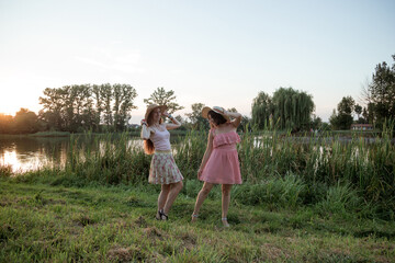 Two young beautiful smiling women in short summer dresses and straw hats posing in front of lake on warm summer day at sunset. Best girlfriends outdoors. Positive models having fun in the park