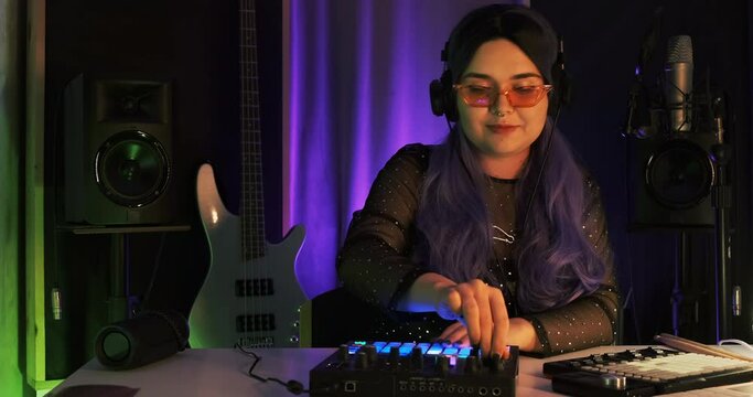 Punk girl model playing live on musical set, electronic music with computer, guitar, launchpad with black headsets and pink grasses. Musician recording DJ mixtape at home studio with violet neon light