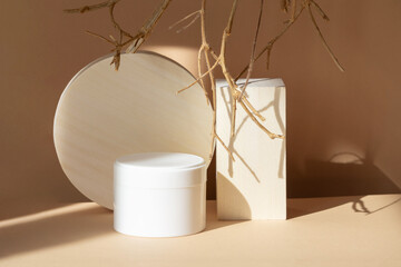 Mock-up of white round jar of body cream next to wooden geometric stands on beige background. Hard shadows from objects fall on surface. Natural tree branches surround 3d podium