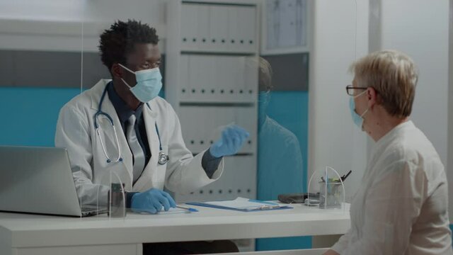 Doctor of african american ethnicity having appointment with elder patient wearing face masks in medical cabinet with plexiglass wall. Black medic consulting caucasian old woman during pandemic