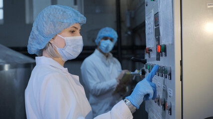 A woman, an employee of an enterprise or a factory, in a sterile white coat, a medical mask, a disposable cap, hands in disposable latex gloves, presses the equipment button, a colleague in the
