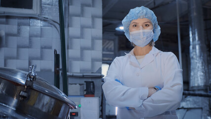 A portrait of a woman, an employee of an enterprise or factory, in a sterile white coat, a medical mask, a disposable cap, hands in disposable latex gloves, posing in front of the camera against the