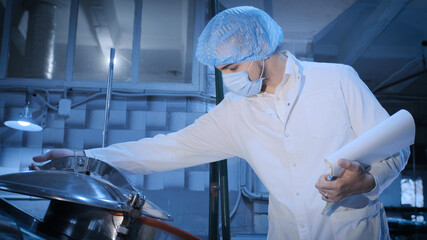 A young process engineer in sterile clothing controls the brewing process of beer or other beverage in a large container in a sterile production hall.