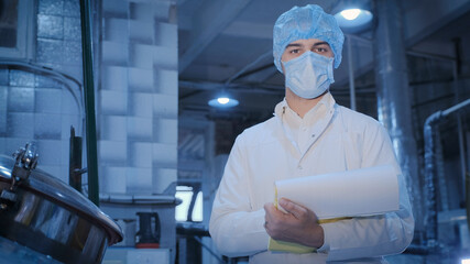 In the production workshop, a young technologist with documents in his hands in a sterile medical mask, disposable cap and in a white medical gown is at a large container.