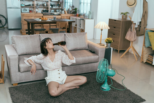 high angle view summer day asian lady going nowhere but staying at home to avoid burning heat outside. taiwanese young female leaning against couch relaxed is guzzling beer alone with electric fan