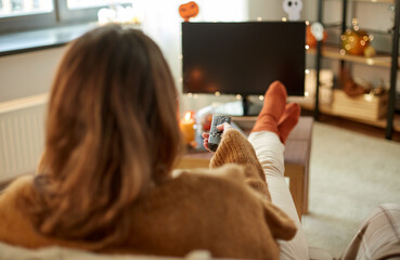 halloween, holidays and leisure concept - young womanwith remote control watching tv and resting her feet on table at cozy home