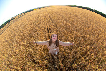 Naklejka na ściany i meble Happy woman stands in a wheat field in the summer against the background of a setting sun