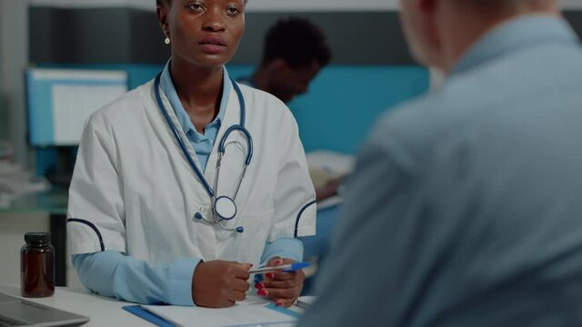 Close up of african american doctor doing consultation with senior patient sitting at desk in medical office. Black medic with white coat giving professional advice to elder man
