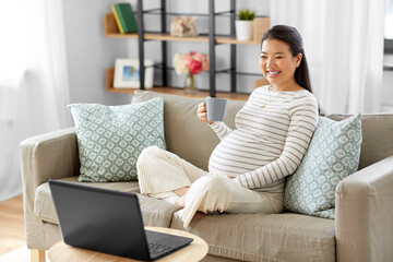 Obraz na płótnie Canvas pregnancy, rest, people and expectation concept - happy smiling pregnant asian woman with laptop computer sitting on sofa at home and drinking tea