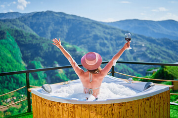 Free carefree girl traveler with open arms relaxing with a glass of wine at hot tube during happy traveling moment vacation life against the background of green big mountains
